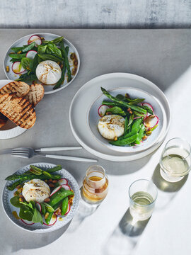 High key still life with glasses of white wine and plates of burrata spring salad on white table, overhead view
