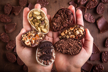 Top view of hands holding six mini stuffed easter eggs on the foreground. Lots of mini easter eggs and cracked chocolates on a wooden table at the background. 