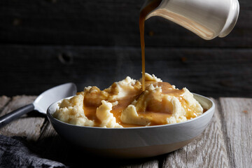 Jug of gravy being poured onto bowl of steaming mashed potatoes, studio shot