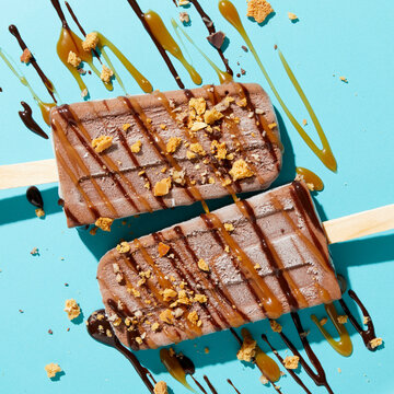 Two homemade fudge iced lollies with sprinkled and drizzled nuts, caramel and chocolate sauce on blue background, overhead view
