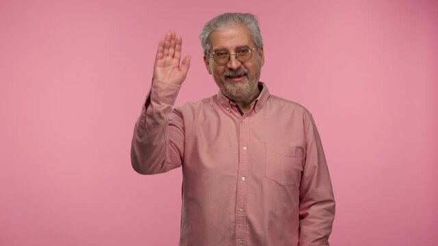 Portrait of an elderly man with glasses waving hand and showing gesture come here. Gray haired pensioner grandfather with beard wearing shirt posing on pink studio background. Slow motion.