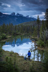 Fototapeta na wymiar A woman sets up her photograph alongside a mirror like reflection in a creek of the mountains in Jasper National Park.