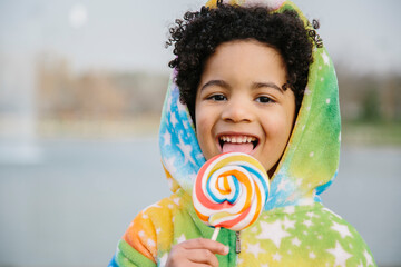 Black boy wearing pajamas, smiling, sucking on a giant lollipop . In a park background. Copyspace. Kids, candies and black people concept