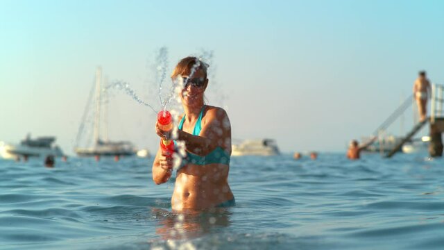 SLOW MOTION, PORTRAIT, CLOSE UP, DOF: Smiling young woman enjoying her summer vacation shoots a water gun at the camera. Fit female tourist is squirting water with plastic toy gun while at the beach.