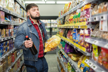 Young bearded man making purchases in supermarket, choosing snacks for beer