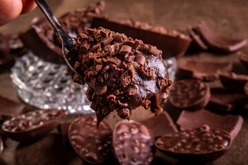 Close up of a spoonful of a cracked stuffed chocolate easter egg with grated chocolate on the top and lots of mini easter eggs and chocolates all around it on a glass stand on a wooden table.