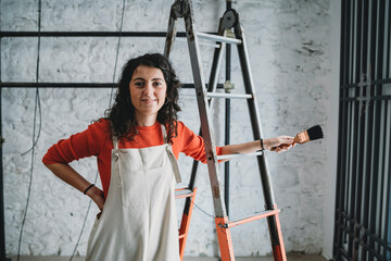 Mid adult woman leaning on step ladder with paint brush in her new shop, portrait