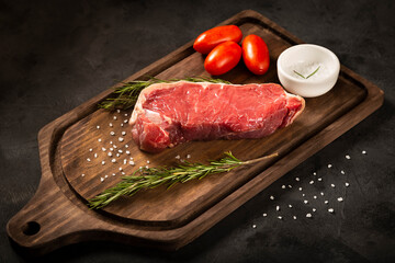 Fresh raw steak on wooden board on a dark background with salt, tomatoes and rosemary.