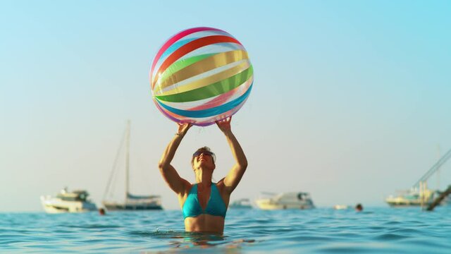 SLOW MOTION, CLOSE UP, DOF: Young woman wearing a blue bikini plays with huge inflatable ball on a sunny summer evening. Female tourist bounces beach ball off her fingertips while playing in ocean