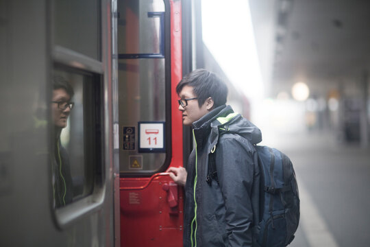 Young Asian man wearing glasses on railway platform, entering passenger train.