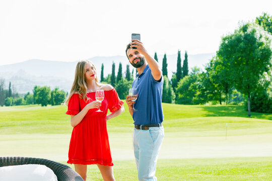 Couple with wine glasses taking selfie on golf course - Powered by Adobe