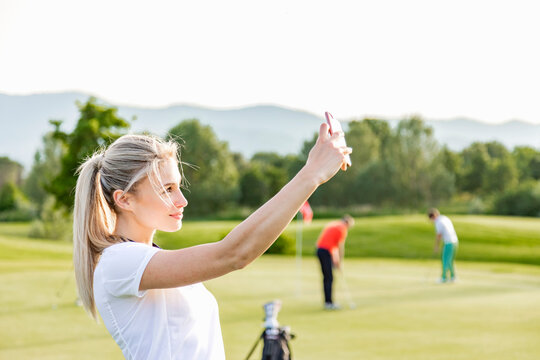 Woman taking selfie on golf course, friends playing golf in background 