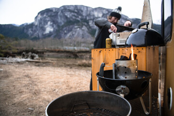 A man cooking on a barbeque set  up at the back of the campervan in a wintry landscape.