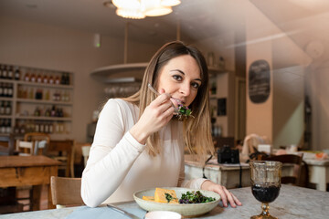 A woman seated at a cafe table eating a dish of vegan food, vegetables on her fork. 