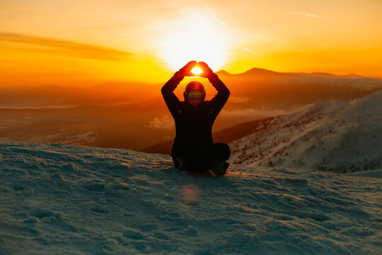 A person sitting on a snow slope, silhouetted against the sunset with arms raised above their head.