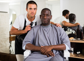 Fototapeta premium Young hairdresser finished haircut of African male client, looking at result together.