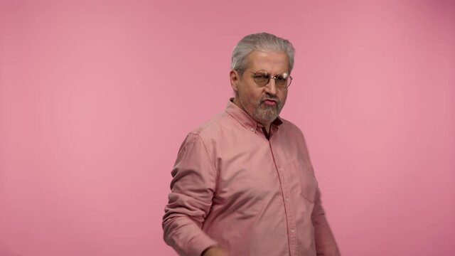 Portrait of an elderly man with glasses making a rock gesture, enjoying life and laughing. Gray haired pensioner grandfather with beard wearing shirt posing on pink studio background. Slow motion.