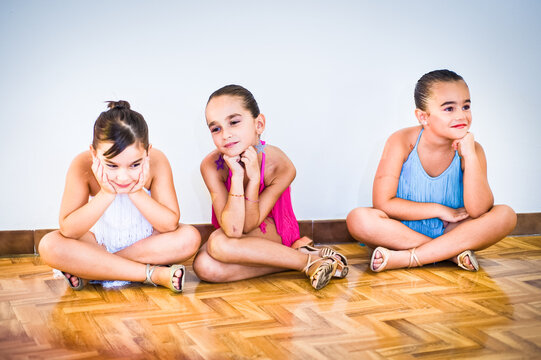 Three young girls at dance class, wearing make up and summer dresses sitting on parquet floor.