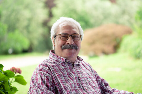 Portrait of senior man with moustache wearing glasses sitting in garden, smiling at camera.