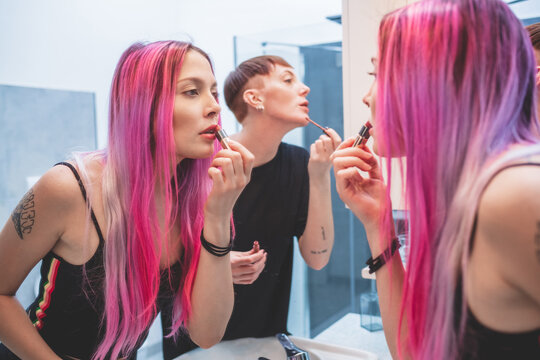 Young woman with long pink hair and woman with short red hair standing in front of mirror, applying lipstick.