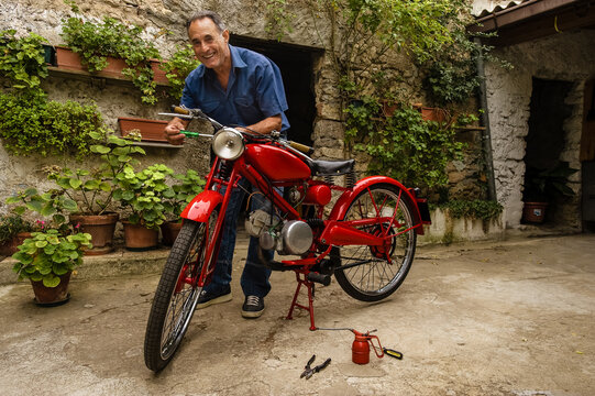 Mature man standing in courtyard, fixing red vintage moorcycle.