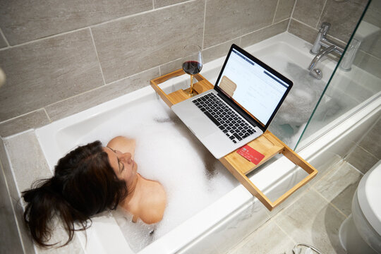 Woman sitting in bathtub, having foam bath and working on laptop during Coronavirus crisis.