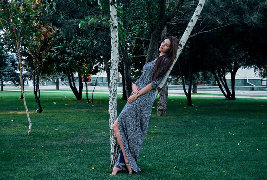 Portrait of woman with long brown hair, wearing long dress, leaning against tree in park.