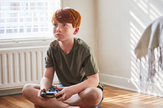 Boy with red hair sitting on floor in sunny room, olding game console controller.