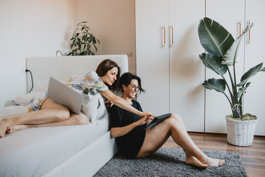 Two women with brown hair sitting on floor and lying on daybed, using laptop and digital tablet.