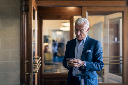Senior businessman standing in hotel lobby, looking at mobile phone. 