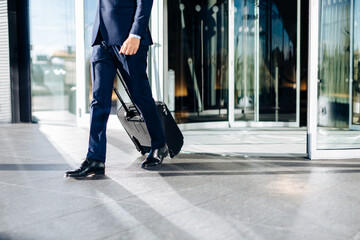 Low section of businessman walking with black trolley through revolving glass door.