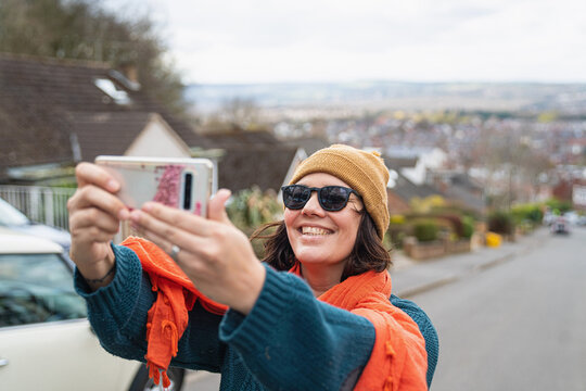 Smiling woman wearing knit hat and sunglasses standing on suburban street, taking selfie with mobile phone.