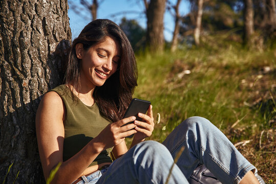 Portrait of young woman with long brown hair sitting under a tree in a forest, checking mobile phone.