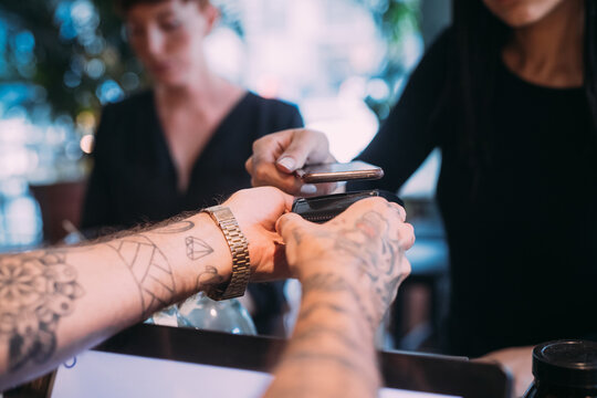 Close up of tattooed bartender standing at counter, handing card reader to woman using mobile phone to pay.