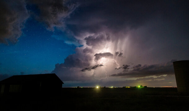 Numerous cloud-to-cloud lightning strikes seen along the dryline at night, with stars in the background over farm buildings