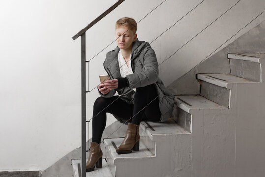 Woman with short blond hair wearing grey puffer jacket sitting on staircase, checking mobile phone.