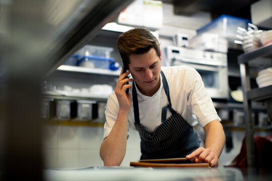Young male chef reading digital tablet and talking on smartphone in kitchen