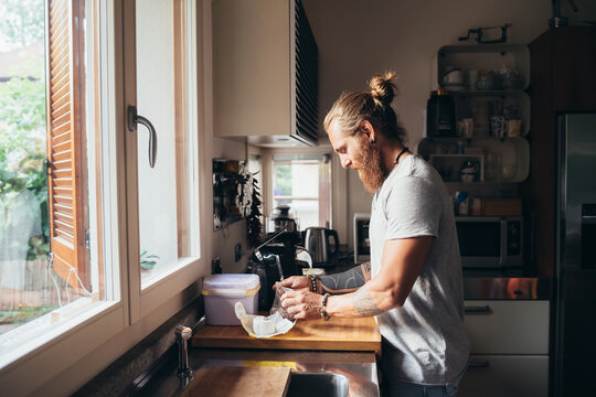 Bearded tattooed man with long brunette hair standing in a kitchen, preparing food.