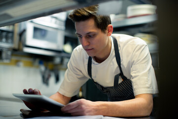 Chef wearing blue apron standing in kitchen, using digital tablet.
