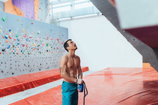 Bare-chested muscular man standing in front of indoor rock climbing wall.