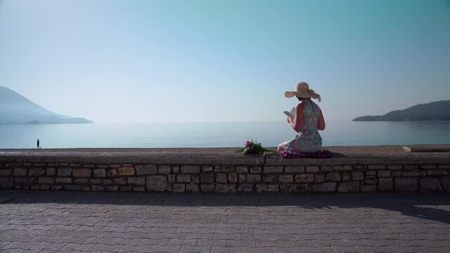 Slim lady in stylish straw hat and colorful dress reads book near bouquet sitting on beach parapet in summer evening backside view