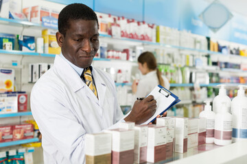 Naklejka na ściany i meble African American man pharmacy specialist making notes on clipboard during inventory in pharmacy