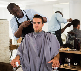 Male client getting trendy haircut at barber shop from African-American hairstylist using machine