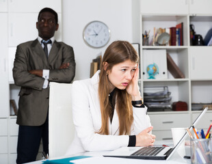 Young woman manager sitting at table after conflict, angry man boss on background