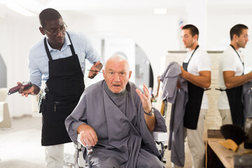 Dissatisfied elderly male client complaining about haircut to perplexed African hairdresser at barber shop