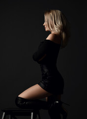 Sexy blonde woman in black leather shorts or mini skirt and boots is standing at stool with her knee on it over dark background. Side view. Fashion, style, sexy wear for women concept - 420934710