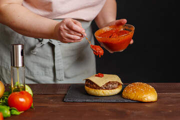 Close-up on the preparation of a magnificent burger. Against the background with ingredients for a burger. Gastronomy, recipes, menus, fast food. Juicy hamburger