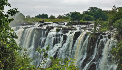 Streams of water cascade down the rocks into the gorge. Splashes and fog over Victoria Falls. Tropical vegetation grows on the banks of the river. Zambia