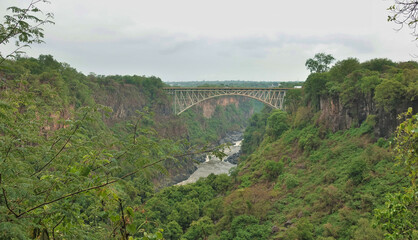The Zambezi River flows in a gorge between steep slopes. A bridge runs over the abyss. In the...