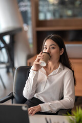 Charming Asian businesswoman drinking coffee and relax at the office. Looking at camera.